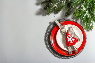 Noel sofrası. Klasik çatal bıçaklı kırmızı seramik tabak, köknar ağacı dalları ve şenlikli aksesuarlar. Modern sert ışık, karanlık gölge. Yaratıcı düz konum, gri arkaplan, üst görünüm