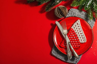 Yılbaşı sofrasını kırmızı tonlarda çatal bıçak setiyle servis etmiş. Evergreen dalları, örülmüş köknar ağaçları, yapay kar. Modern sert ışık, karanlık gölge. Düz konum, kırmızı arkaplan, üst görünüm