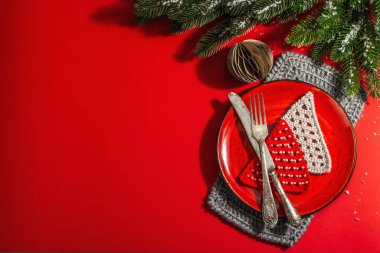 Yılbaşı sofrasını kırmızı tonlarda çatal bıçak setiyle servis etmiş. Evergreen dalları, örülmüş köknar ağaçları, yapay kar. Modern sert ışık, karanlık gölge. Düz konum, kırmızı arkaplan, üst görünüm