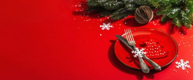 Yılbaşı sofrasını kırmızı tonlarda çatal bıçak setiyle servis etmiş. Evergreen dalları, örülmüş köknar ağaçları, yapay kar. Modern sert ışık, karanlık gölge. Düz konum, kırmızı arkaplan, pankart biçimi