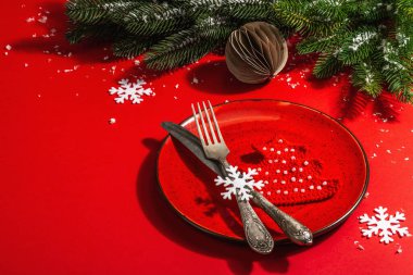 Yılbaşı sofrasını kırmızı tonlarda çatal bıçak setiyle servis etmiş. Evergreen dalları, örülmüş köknar ağaçları, yapay kar. Modern sert ışık, karanlık gölge. Düz konum, kırmızı arkaplan, kopyalama alanı