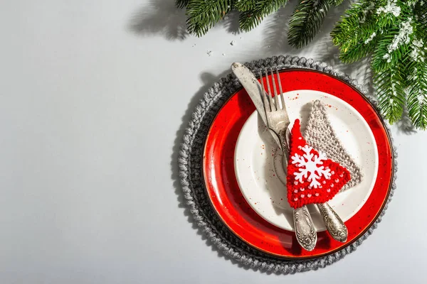 Noel sofrası. Klasik çatal bıçaklı kırmızı seramik tabak, köknar ağacı dalları ve şenlikli aksesuarlar. Modern sert ışık, karanlık gölge. Yaratıcı düz konum, gri arkaplan, üst görünüm
