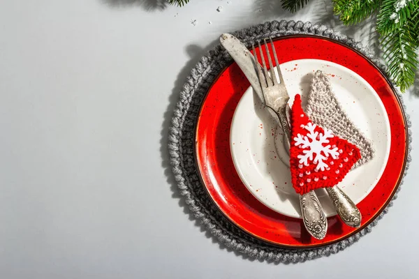 Noel sofrası. Klasik çatal bıçaklı kırmızı seramik tabak, köknar ağacı dalları ve şenlikli aksesuarlar. Modern sert ışık, karanlık gölge. Yaratıcı düz konum, gri arkaplan, üst görünüm