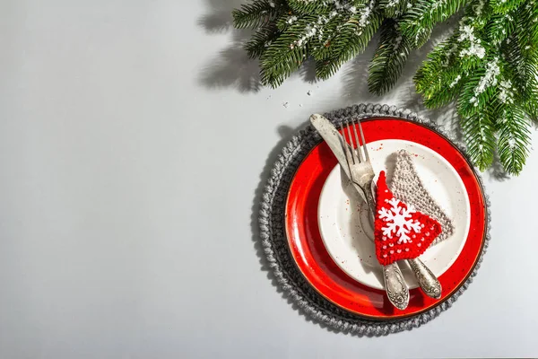 Noel sofrası. Klasik çatal bıçaklı kırmızı seramik tabak, köknar ağacı dalları ve şenlikli aksesuarlar. Modern sert ışık, karanlık gölge. Yaratıcı düz konum, gri arkaplan, üst görünüm