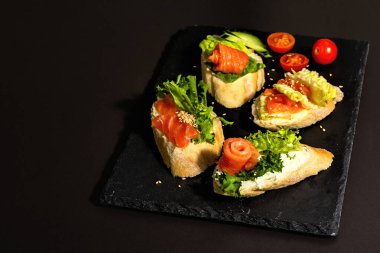 Assorted open sandwiches with salted salmon, cream cheese, salad leaves and cucumber. Seafood, healthy food. Black stone concrete background, hard light, dark shadow, copy space