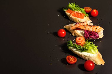 Assorted open sandwiches with salted salmon, shrimps, cream cheese, salad leaves and cucumber. Seafood, healthy food. Black stone concrete background, hard light, dark shadow, copy space
