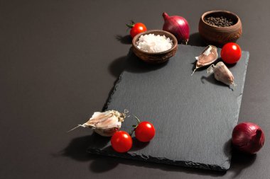 Yemek pişirme konsepti. Seramik standı, domates kirazı, sarımsak, deniz tuzu, biber ve mor soğan. Modaya uygun bir sert ışık, karanlık gölge, siyah taş beton zemin, kopyalama alanı