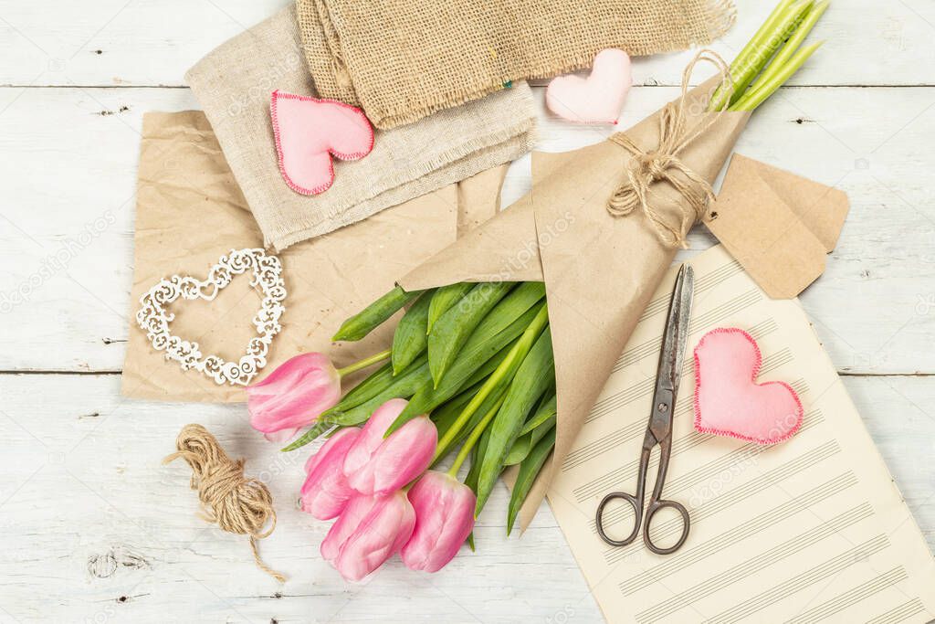 Bouquet of fresh pink tulips. Making spring floral decorations. Tools, vintage scissors, craft paper, soft hearts. White wooden boards background, top view