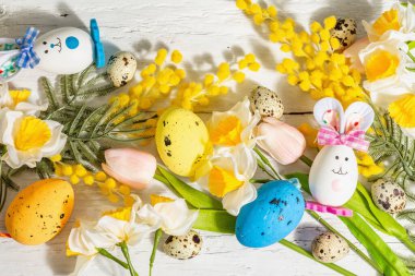 Pastel yumurtalar, sevimli tavşanlar ve geleneksel bahar çiçekleri olan Paskalya bayramı arkaplanı. Sert ışık, karanlık gölge, beyaz ahşap zemin, üst manzara