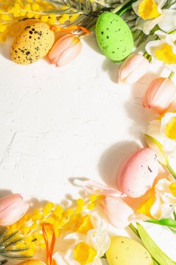 Pastel yumurtalar ve geleneksel bahar çiçekleriyle bahar bayramı arkaplanı. Sert ışık, karanlık gölge, beyaz ahşap zemin, üst manzara