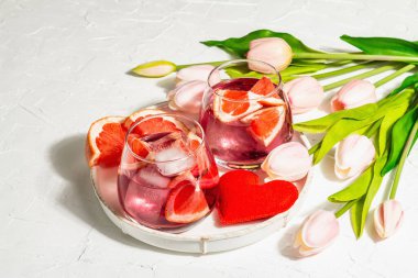 Greyfurtlu alkolsüz kokteyl. Bardakta buzlu içecek servis edilir. Sevgililer Günü sembolü bir kalp ve nazik lalelerdir. Sert ışık, karanlık gölge, beyaz ahşap zemin, kapat