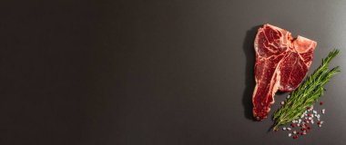 Siyah beton arka planda baharatlı klasik T-bone bifteği. Taze biberiye, modaya uygun sert ışık, koyu gölge, düz yer, üst manzara, pankart biçimi