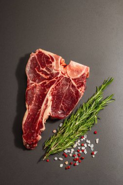Siyah beton arka planda baharatlı klasik T-bone bifteği. Taze biberiye, modaya uygun sert ışık, koyu gölge, düz yer, üst manzara