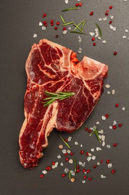 Siyah beton arka planda baharatlı klasik T-bone bifteği. Taze biberiye, çatal bıçak takımı, modaya uygun sert ışık, koyu gölge, düz yer, üst manzara
