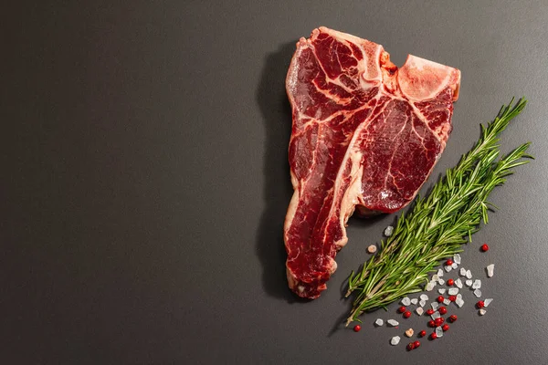 Siyah beton arka planda baharatlı klasik T-bone bifteği. Taze biberiye, modaya uygun sert ışık, koyu gölge, düz yer, üst manzara