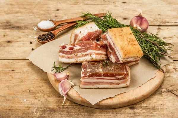 Pieces of salty high-fat meat cooked with spices. Salo, bacon, lard, silverside, gammon. Garlic, fresh rosemary, spices. Hard light, dark shadow, wooden background, copy space