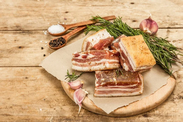 Pieces of salty high-fat meat cooked with spices. Salo, bacon, lard, silverside, gammon. Garlic, fresh rosemary, spices. Hard light, dark shadow, wooden background, copy space