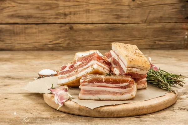 Pieces of salty high-fat meat cooked with spices. Salo, bacon, lard, silverside, gammon. Garlic, fresh rosemary, spices. Hard light, dark shadow, wooden background, copy space