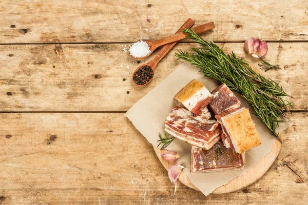 Pieces of salty high-fat meat cooked with spices. Salo, bacon, lard, silverside, gammon. Garlic, fresh rosemary, spices. Hard light, dark shadow, wooden background, top view