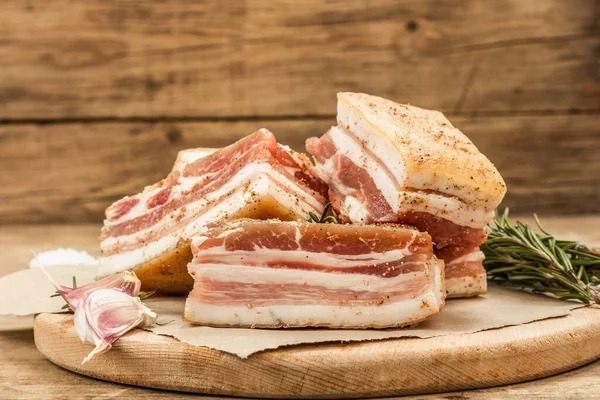 Pieces of salty high-fat meat cooked with spices. Salo, bacon, lard, silverside, gammon. Garlic, fresh rosemary, spices. Hard light, dark shadow, wooden background, copy space