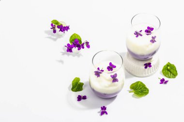Taze menekşe çiçekli vegan panna cotta tatlısı. Çizgili renkli yiyecekler, modaya uygun stantlar. Sert bir ışık, koyu gölge, beyaz arkaplan, kopyalama alanı