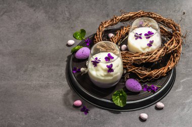 Mutlu Paskalya bayramı kompozisyonu. Taze menekşe çiçekli vegan panna cotta tatlısı. Çizgili renkli yiyecekler, modaya uygun stantlar. Sert bir ışık, karanlık bir gölge, taş beton zemin, kopyalama alanı