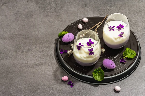 Mutlu Paskalya bayramı kompozisyonu. Taze menekşe çiçekli vegan panna cotta tatlısı. Çizgili renkli yiyecekler, modaya uygun stantlar. Sert bir ışık, karanlık bir gölge, taş beton zemin, kopyalama alanı