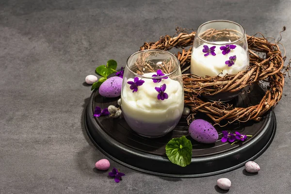Mutlu Paskalya bayramı kompozisyonu. Taze menekşe çiçekli vegan panna cotta tatlısı. Çizgili renkli yiyecekler, modaya uygun stantlar. Sert bir ışık, karanlık bir gölge, taş beton zemin, kopyalama alanı