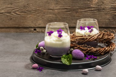 Mutlu Paskalya bayramı kompozisyonu. Taze menekşe çiçekli vegan panna cotta tatlısı. Çizgili renkli yiyecekler, modaya uygun stantlar. Sert bir ışık, karanlık bir gölge, taş beton zemin, kopyalama alanı