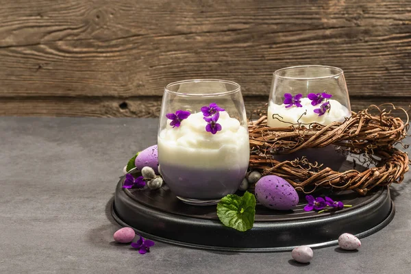 Mutlu Paskalya bayramı kompozisyonu. Taze menekşe çiçekli vegan panna cotta tatlısı. Çizgili renkli yiyecekler, modaya uygun stantlar. Sert bir ışık, karanlık bir gölge, taş beton zemin, kopyalama alanı