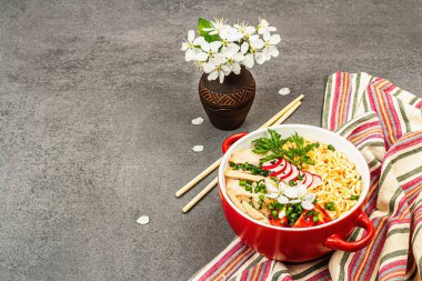 Kırmızı bir kasede tavuk, sebze ve otlu Asya eriştesi. Geleneksel Japon çorbası, çorba ve kiraz çiçekleri. Gri taş beton zemin, kopyalama alanı