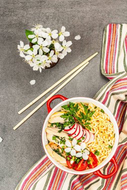 Kırmızı bir kasede tavuk, sebze ve otlu Asya eriştesi. Geleneksel Japon çorbası, çorba ve kiraz çiçekleri. Gri taş arkaplan, üst manzara