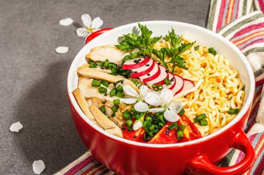 Kırmızı bir kasede tavuk, sebze ve otlu Asya eriştesi. Geleneksel Japon çorbası, çorba ve kiraz çiçekleri. Taş beton arka plan, modaya uygun sert ışık, karanlık gölge, kapat