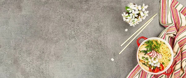 Kırmızı bir kasede tavuk, sebze ve otlu Asya eriştesi. Geleneksel Japon çorbası, çorba ve kiraz çiçekleri. Gri taş arkaplan, pankart biçimi