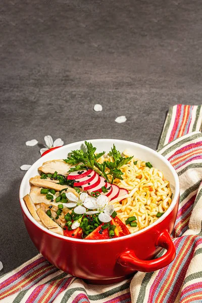 Kırmızı bir kasede tavuk, sebze ve otlu Asya eriştesi. Geleneksel Japon çorbası, çorba ve kiraz çiçekleri. Taş beton zemin, modaya uygun sert ışık, karanlık gölge, kopyalama alanı