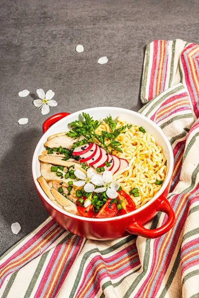 Kırmızı bir kasede tavuk, sebze ve otlu Asya eriştesi. Geleneksel Japon çorbası, çorba ve kiraz çiçekleri. Taş beton arka plan, modaya uygun sert ışık, karanlık gölge, kapat