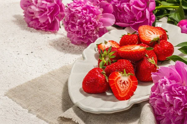 Sweet ripe strawberries and fragrant peonies bouquet. Summer plaster background, seasonal design, hard light, dark shadow, copy space
