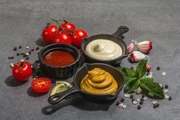 stock image Mustard, ketchup and mayonnaise sauces. Fresh mint, lemon, garlic, and spices. A hard light, dark shadow, stone concrete background, flat lay, close up