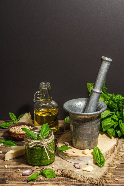 Homemade Italian basil pesto sauce in a vintage mortar with pestle. Fresh bunch of leaves, parmesan, pine nuts, and olive oil. Trendy hard light, dark shadow, old wooden background, copy space