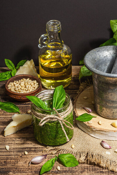 Homemade Italian basil pesto sauce in a vintage mortar with pestle. Fresh bunch of leaves, parmesan, pine nuts, and olive oil. Trendy hard light, dark shadow, old wooden background, close up