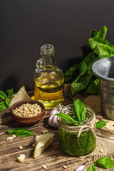 Homemade Italian basil pesto sauce in a vintage mortar with pestle. Fresh bunch of leaves, parmesan, pine nuts, and olive oil. Trendy hard light, dark shadow, old wooden background, close up