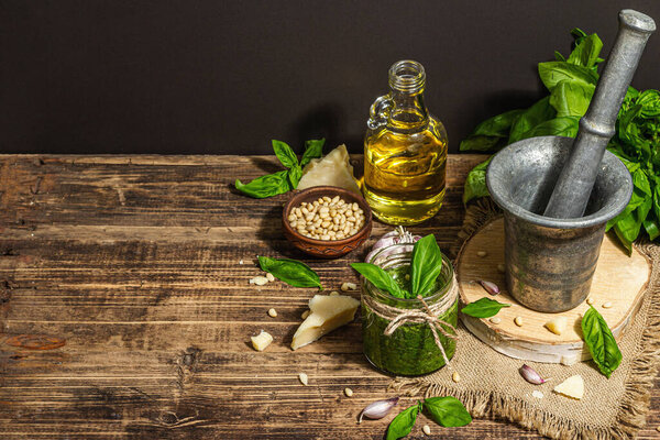 Homemade Italian basil pesto sauce in a vintage mortar with pestle. Fresh bunch of leaves, parmesan, pine nuts, and olive oil. Trendy hard light, dark shadow, old wooden background, copy space