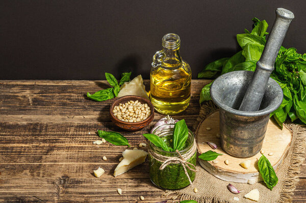 Homemade Italian basil pesto sauce in a vintage mortar with pestle. Fresh bunch of leaves, parmesan, pine nuts, and olive oil. Trendy hard light, dark shadow, old wooden background, copy space