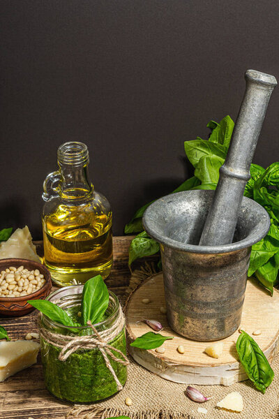 Homemade Italian basil pesto sauce in a vintage mortar with pestle. Fresh bunch of leaves, parmesan, pine nuts, and olive oil. Trendy hard light, dark shadow, old wooden background, copy space