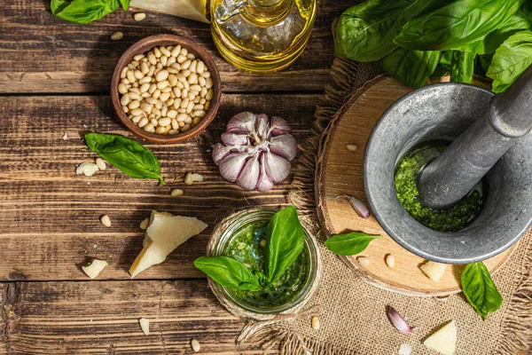 Homemade Italian basil pesto sauce in a vintage mortar with pestle. Fresh bunch of leaves, parmesan, pine nuts, and olive oil. Trendy hard light, dark shadow, old wooden background, top view