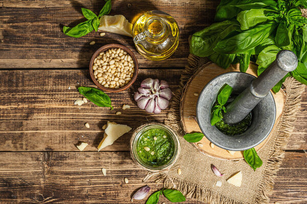Homemade Italian basil pesto sauce in a vintage mortar with pestle. Fresh bunch of leaves, parmesan, pine nuts, and olive oil. Trendy hard light, dark shadow, old wooden background, top view