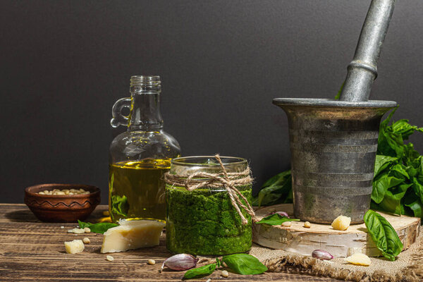 Homemade Italian basil pesto sauce in a vintage mortar with pestle. Fresh bunch of leaves, parmesan, pine nuts, and olive oil. Trendy hard light, dark shadow, old wooden background, copy space