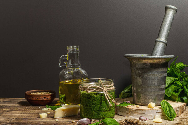 Homemade Italian basil pesto sauce in a vintage mortar with pestle. Fresh bunch of leaves, parmesan, pine nuts, and olive oil. Trendy hard light, dark shadow, old wooden background, copy space