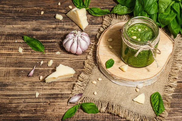 Homemade Italian basil pesto sauce in a glass jar. Fresh bunch of leaves, parmesan, pine nuts, and olive oil. Trendy hard light, dark shadow, old wooden background, copy space