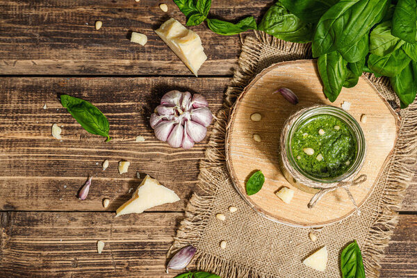 Homemade Italian basil pesto sauce in a glass jar. Fresh bunch of leaves, parmesan, pine nuts, and olive oil. Trendy hard light, dark shadow, old wooden background, top view
